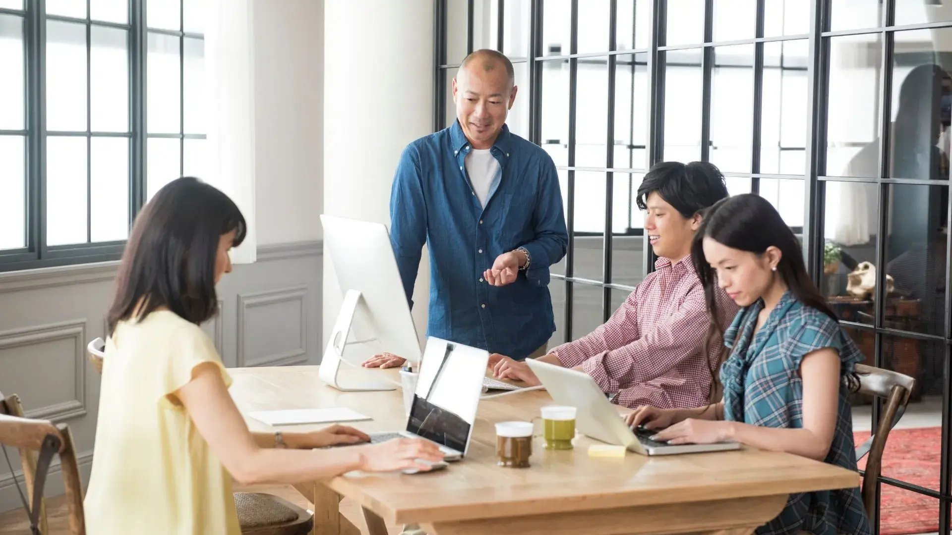 A group of four officemates are working together while facing their laptop screens. One of them seems to be explaining something while looking at one of the laptop screens. 