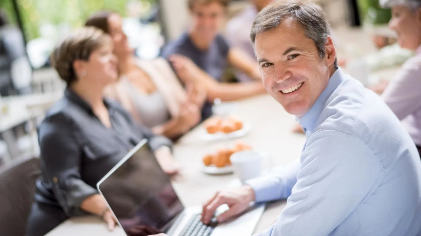 In the foreground, a male office worker is smiling at the camera while his laptop is open. In the background are his colleagues presumably in the middle of a meeting.