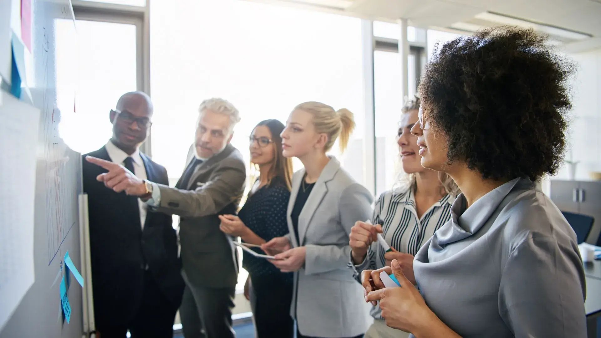 A team of corporate workers are gathered around a whiteboard filled with written notes. One of them is pointing to something on the board while the others are listening intently. 