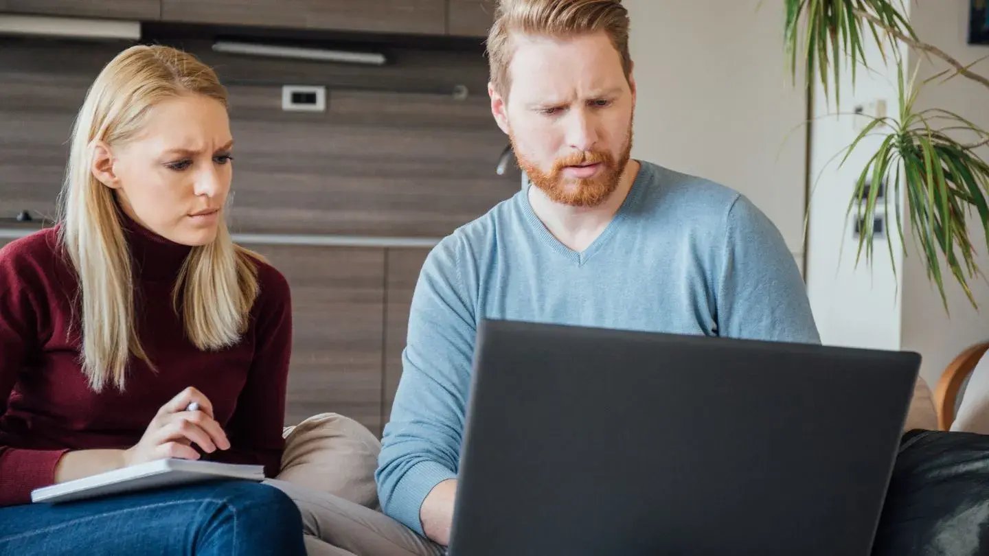 A male office worker has his forehead creased while looking at his laptop. His female colleague also looks frustrated as she stares at his laptop screen.