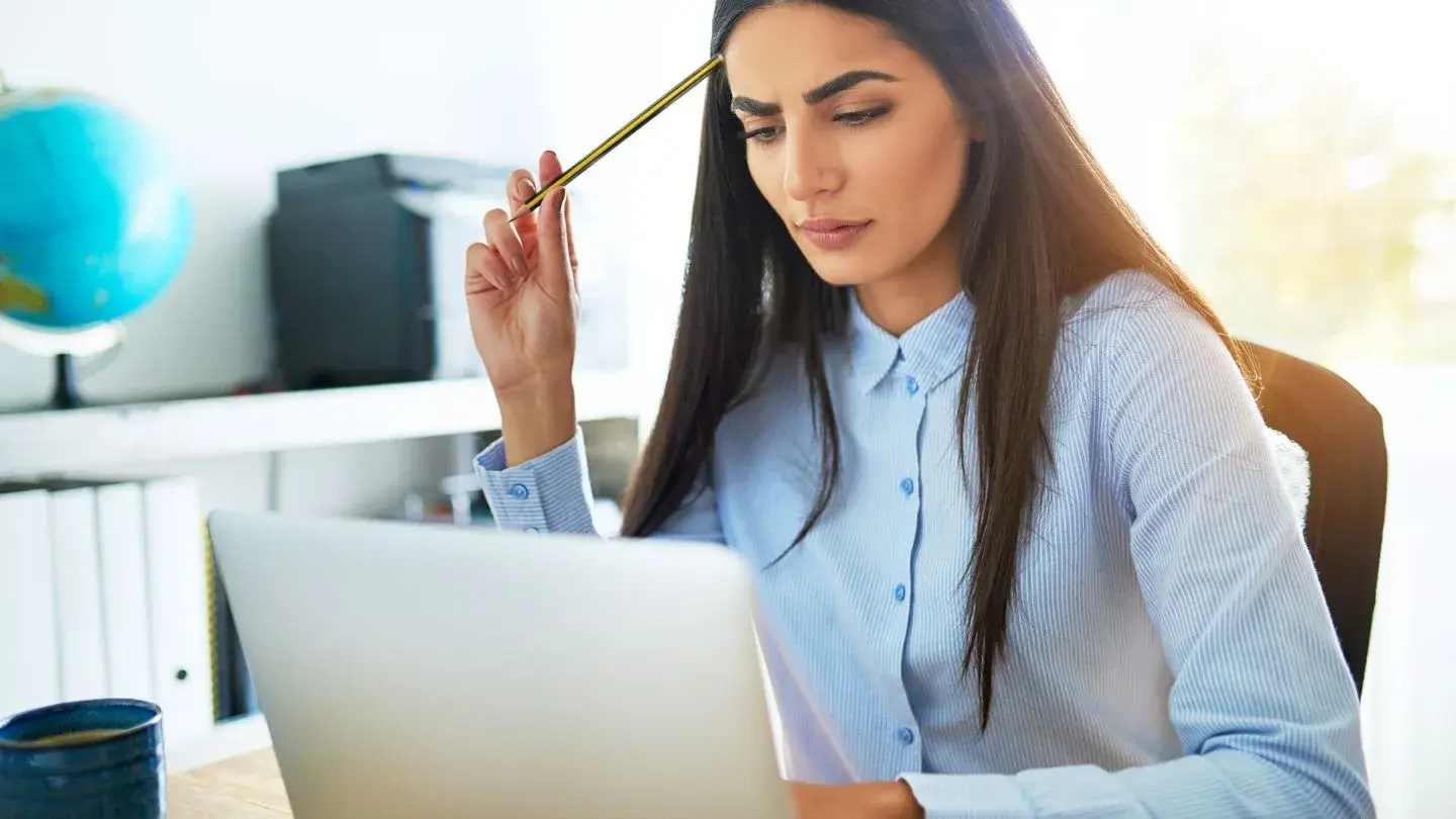 A female Indian office worker is frowning while holding a pencil to her forehead and looking at her laptop screen.